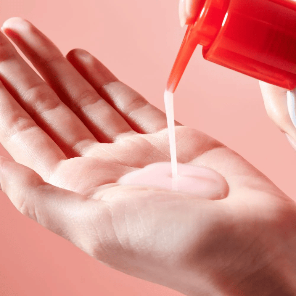 Een lichtroze reinigingsolie wordt aangebracht vanuit een rode pompflacon in een open hand tegen een roze achtergrond.