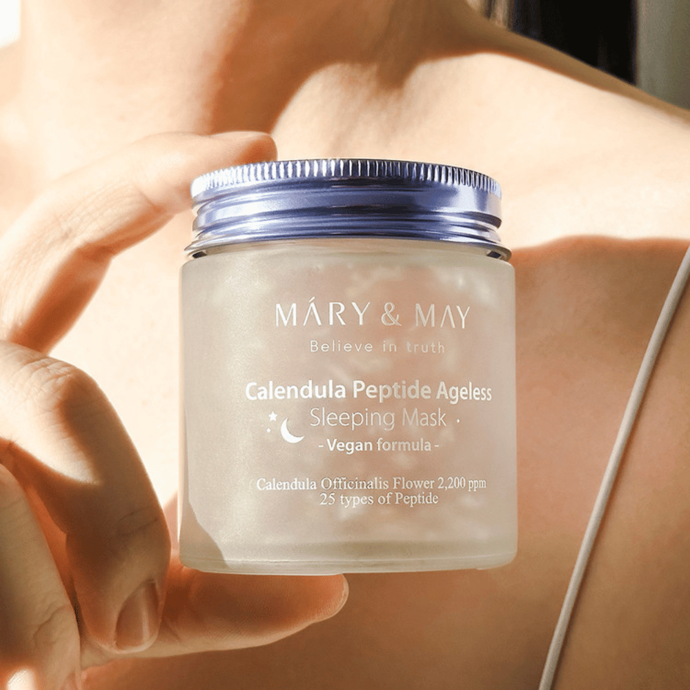 Een vrouw houdt een pot met MARY & MAY Calendula Peptide Ageless Sleeping Mask vast. De pot is transparant met een paarse dop en de tekst "Calendula Peptide Ageless Sleeping Mask" is duidelijk zichtbaar. Het product is een veganistische slaapmasker met calendula en peptiden, bedoeld om de huid 's nachts een jeugdige glans te geven.