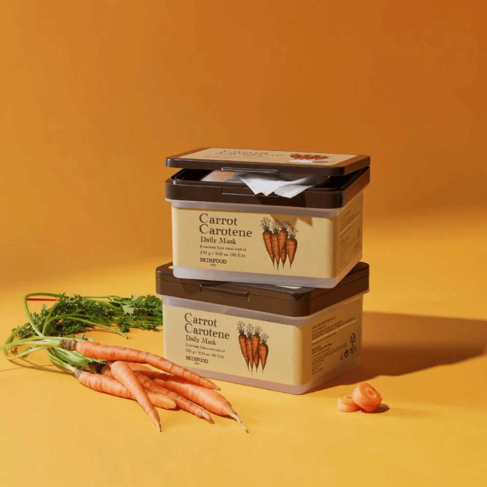 De afbeelding toont drie gestapelde verpakkingen van "Carrot Carotene Daily Mask" van SKINFOOD, geplaatst op een oranje achtergrond samen met verse wortels. Elke doos bevat gezichtsmaskers verrijkt met β-caroteen uit wortelzaadolie, ontworpen voor dagelijks gebruik om de huid te voeden en te verzorgen.