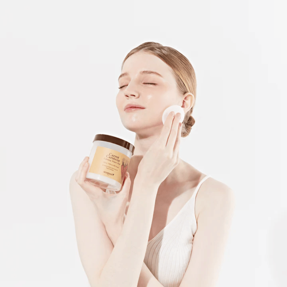 Een vrouw met opgestoken haar brengt een huidverzorgingsproduct aan op haar gezicht met een wattenschijfje. Ze houdt een grote pot vast gemarkeerd met "Carrot Carotene Calming Water Pad". Haar gesloten ogen en vredige uitdrukking geven een gevoel van ontspanning en plezier in de huidverzorgingsroutine.