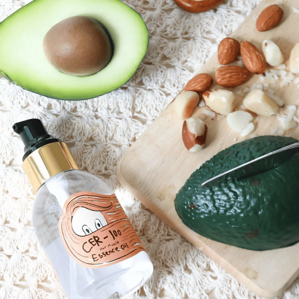 Elizavecca CER-100 Hair Muscle Essence Oil ingrediënten – close-up met avocado en plantaardige oliën die een voedende haarolie tonen die helpt het haar te verzachten en te verzorgen.