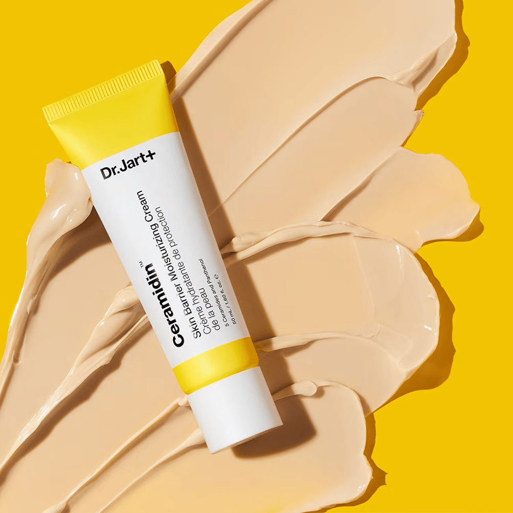 Tube met Dr. Jart+ Ceramidin Skin Barrier Moisturizing Cream tegen een gele achtergrond met verspreide crème.