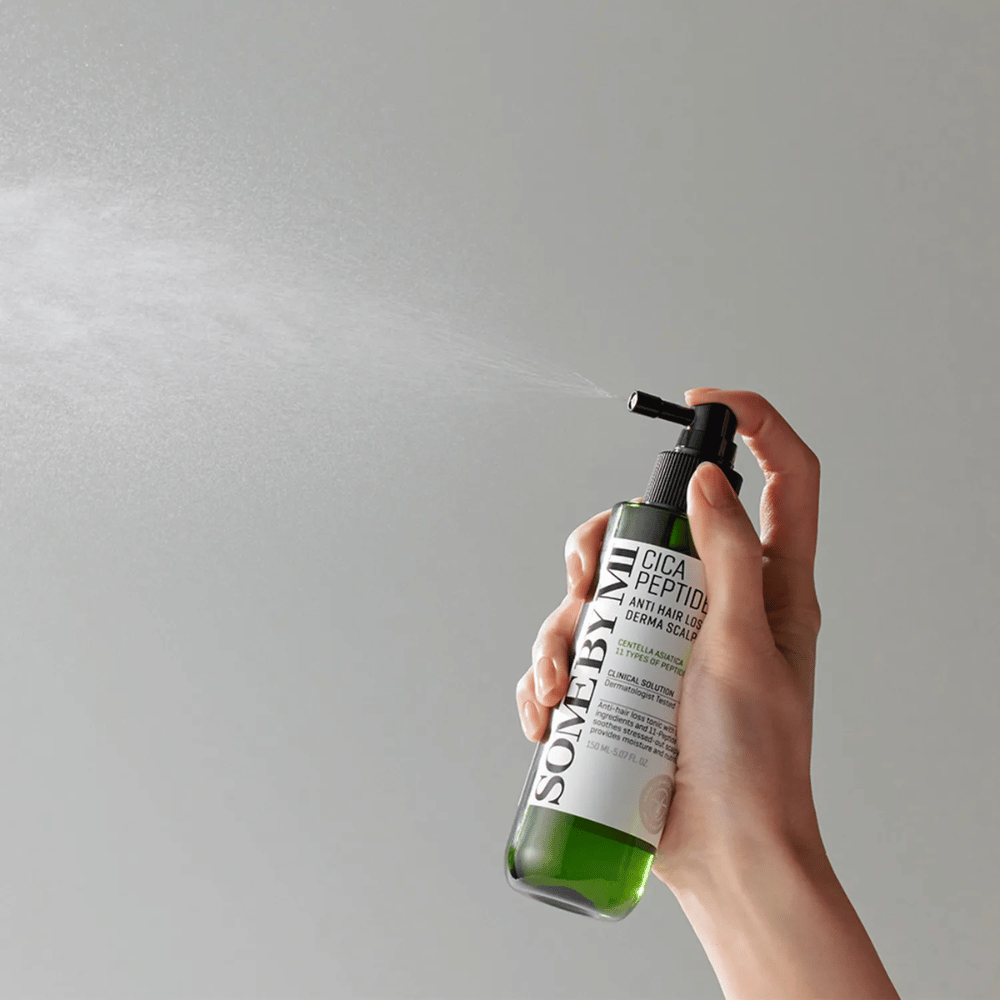 Hand die SOME BY MI Cica Peptide Anti Hair Loss Derma Scalp Tonic spuit uit een groene fles met zwart mondstuk en wit etiket tegen een grijze achtergrond.