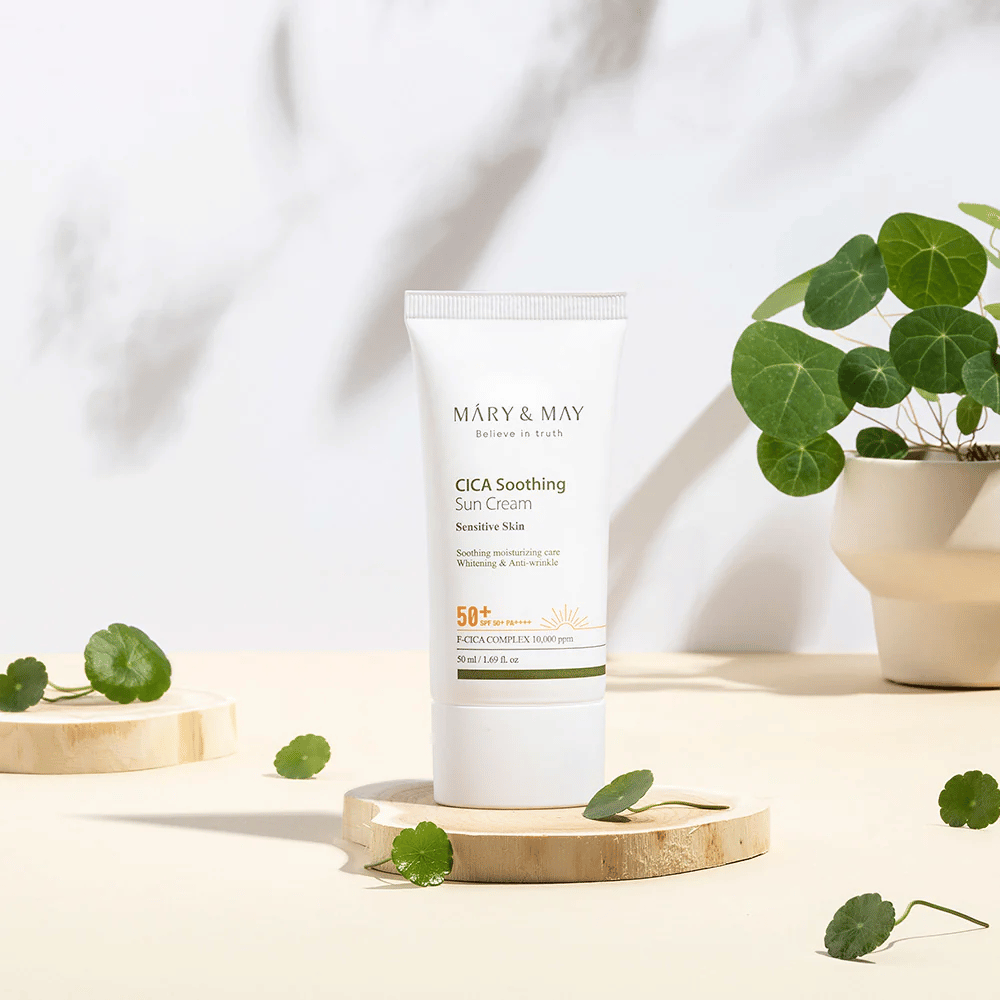 Mary&May CICA Soothing Sun Cream geplaatst op een houten podium met groene centella-bladeren eromheen. De afbeelding benadrukt de huidkalmerende ingrediënten en de natuurlijke inspiratie achter het product.