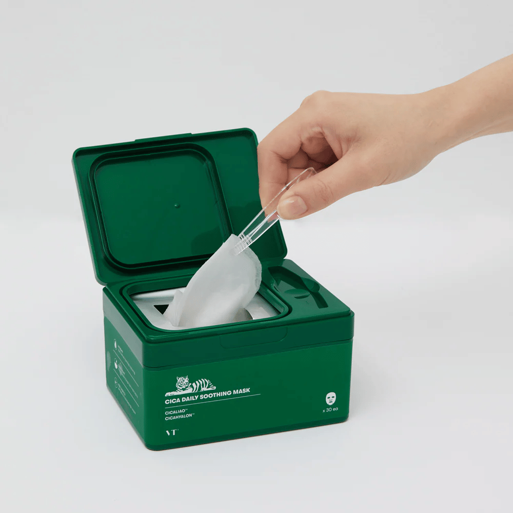 Hand gebruikt een pincet om een Sheet mask uit een groene houder gemarkeerd "Cica Daily Soothing Mask" te halen.