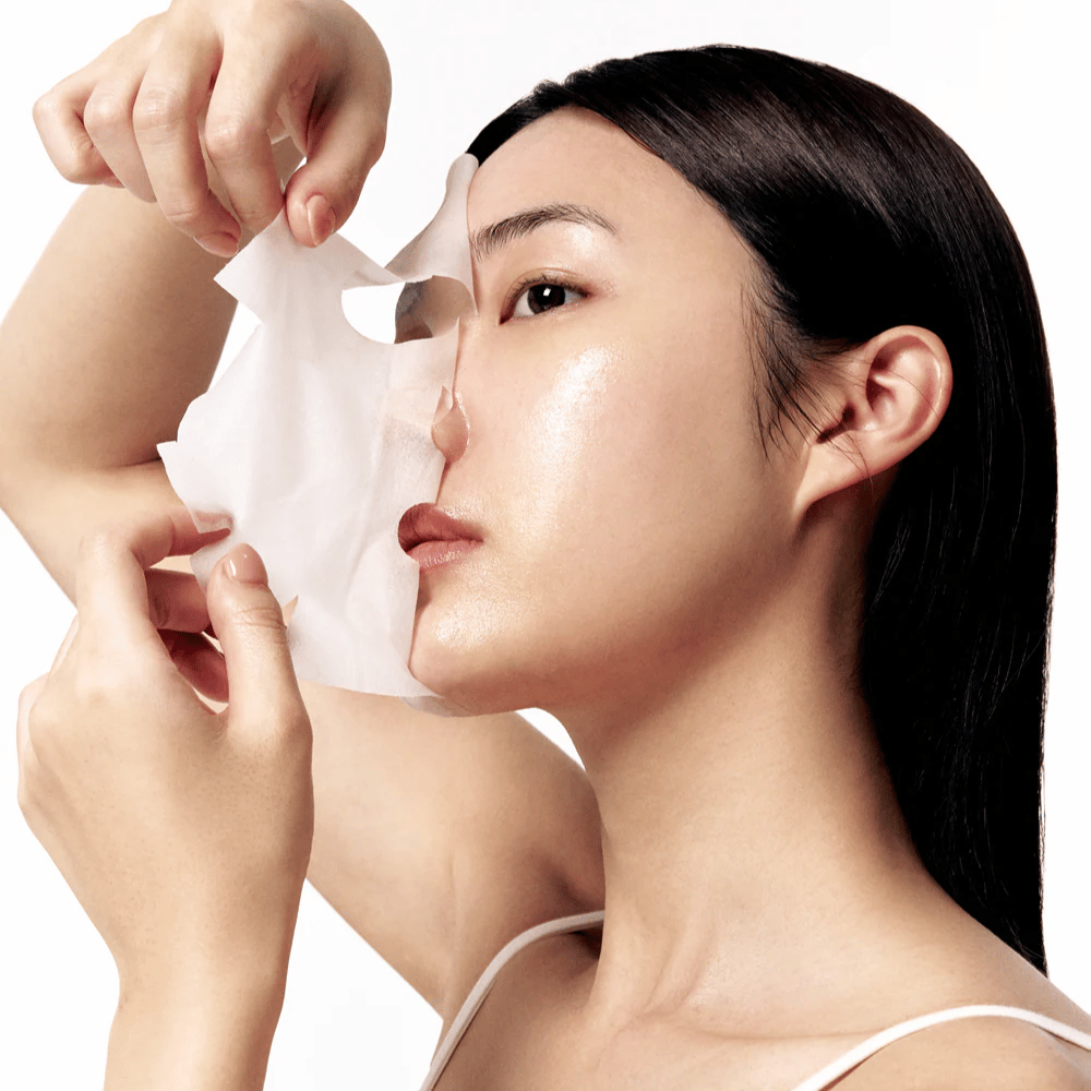 Vrouw met gladde, gehydrateerde huid haalt een witte Sheet mask van het gezicht, wat een stralende en egale huidtoon onthult.
