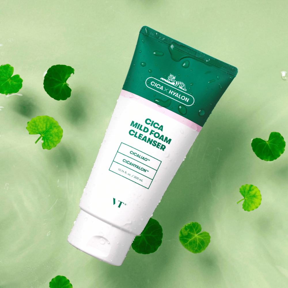 VT Cica Mild Foam Cleanser liggend op een lichtgroene ondergrond met waterdruppels op de verpakking en omgeven door groene centella asiatica-bladeren, wat de kalmerende eigenschappen van het product benadrukt.