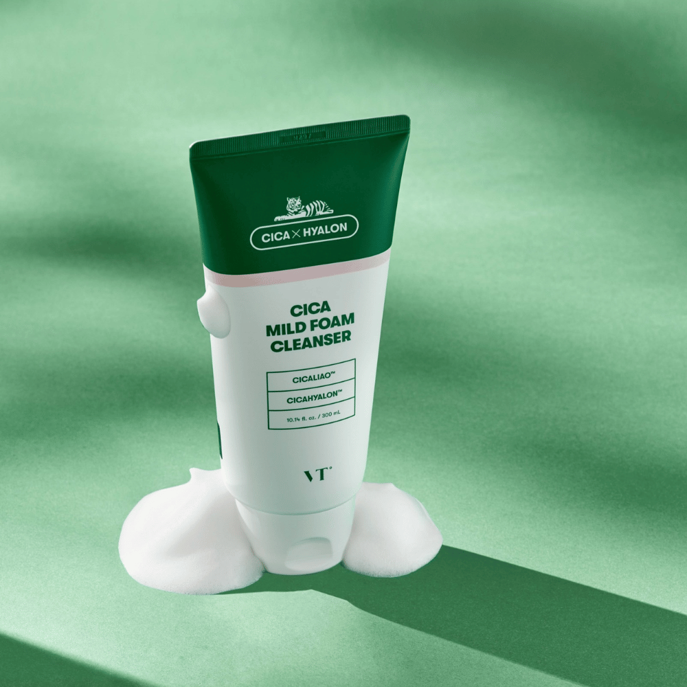 Tube met VT Cica Mild Foam Cleanser staand op een groene ondergrond met schuim rond de basis. Het product heeft een groen en wit ontwerp en wordt gepromoot met CICALIAO™ en CICAHYALON™.