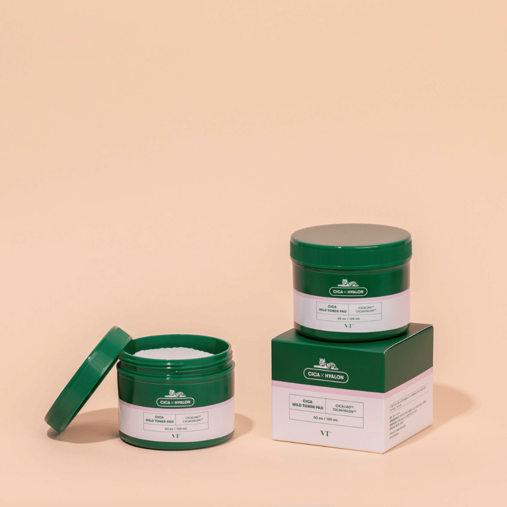 Twee groene potten met VT Cica Mild Toner Pad, een open die de witte toner pads toont en een gesloten geplaatst bovenop zijn productverpakking, tegen een beige achtergrond.