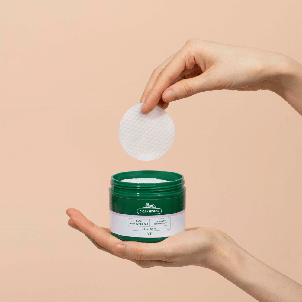 Hand die een groene pot met VT Cica Mild Toner Pad vasthoudt en een ronde, witte toner pad boven de pot optilt tegen een beige achtergrond.