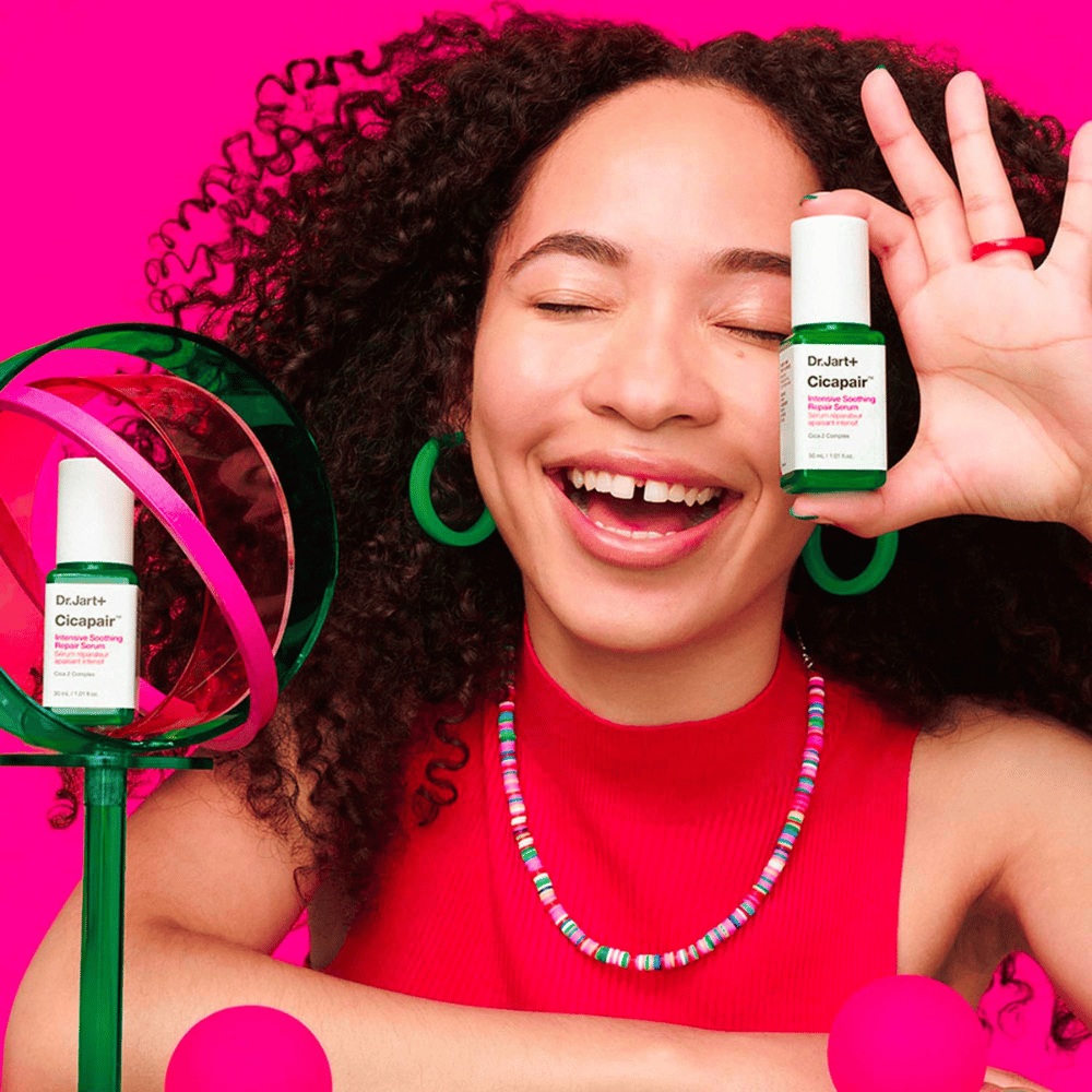 Een vrouw lacht breed terwijl ze Dr.Jart+ Cicapair Intensive Soothing Repair Serum voor haar gezicht houdt. Ze draagt groene oorbellen en een rode ketting. De achtergrond is lichtroze en versierd met groene en roze cirkels.