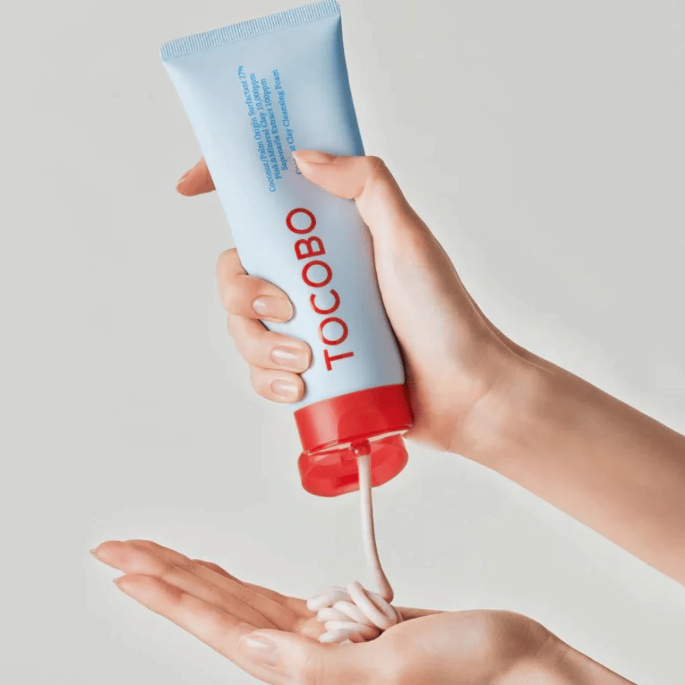 Een persoon houdt een tube van TOCOBO Coconut Clay Cleansing Foam vast. De tube is lichtblauw met een rode dop en de tekst "TOCOBO" aan de voorkant. Het product wordt uit de tube geperst en vormt zich als een shampoo in de hand. De achtergrond is neutraal en legt de focus op het product.