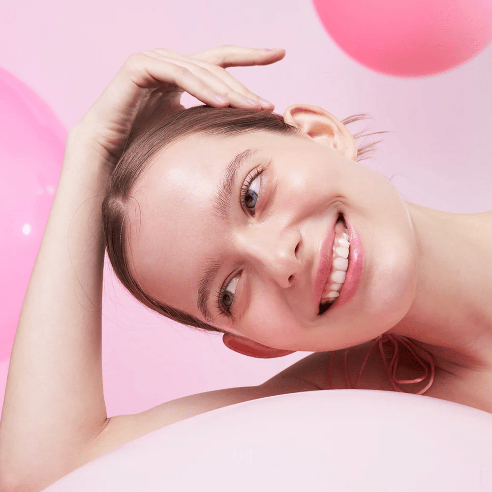 Een vrouw glimlacht en rust haar hoofd op haar hand, omringd door roze luchtballonnen. Haar huid ziet er stralend en natuurlijk gezond uit.