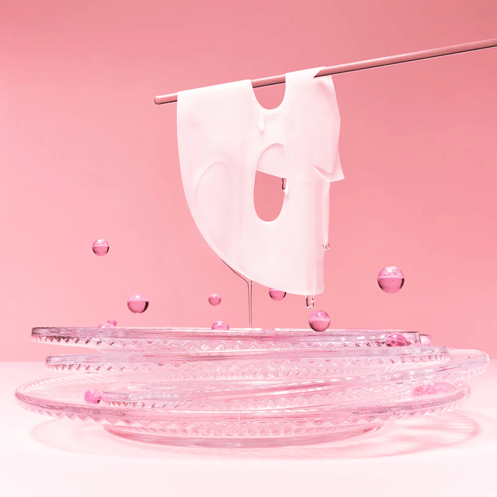 Een hydrogellaagmasker hangt aan een staaf, druipend van het serum. De achtergrond is roze en de spiegel aan de onderkant weerspiegelt de roze bellen die het masker omringen.