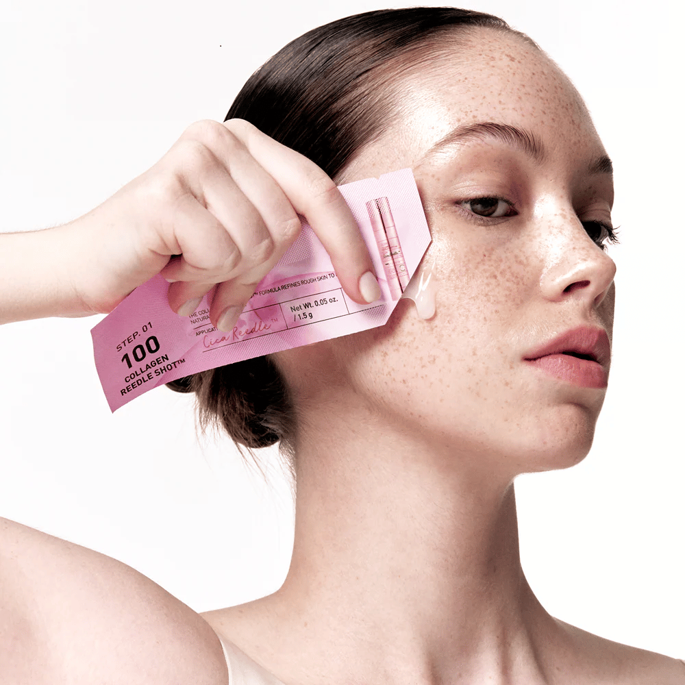 Vrouw brengt stap 1 van Collagen Reedle Shot 100 aan met een roze wegwerpverpakking op de wang. Een heldere gel is zichtbaar op de huid, wat de eerste stap in de tweestaps huidverzorgingsbehandeling markeert.