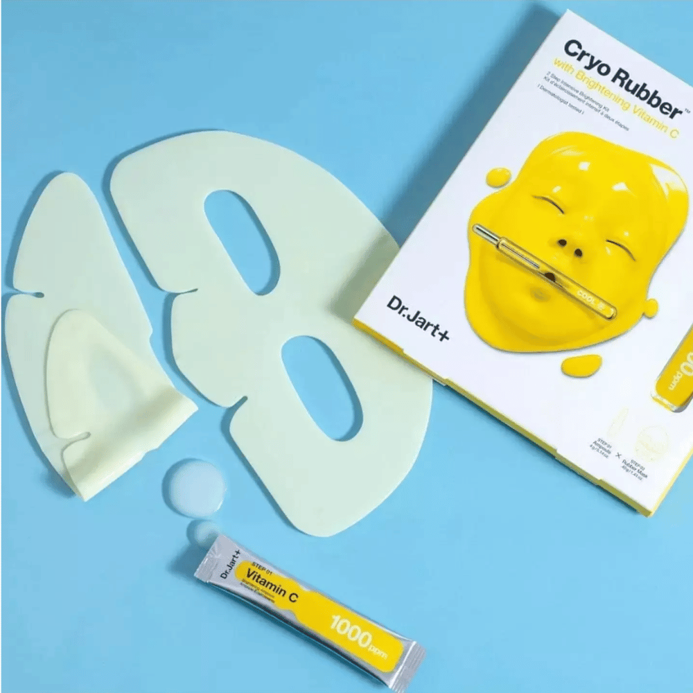 Afbeelding van Dr.Jart+'s 'Cryo Rubber™ met Brightening Vitamin C' schoonheidsproduct. Op een blauwe achtergrond is een wit rubberen gezichtsmasker te zien, een kleine tube met het opschrift 'Vitamine C 1000ppm', en een doos met de productnaam en afbeelding van het gele masker.