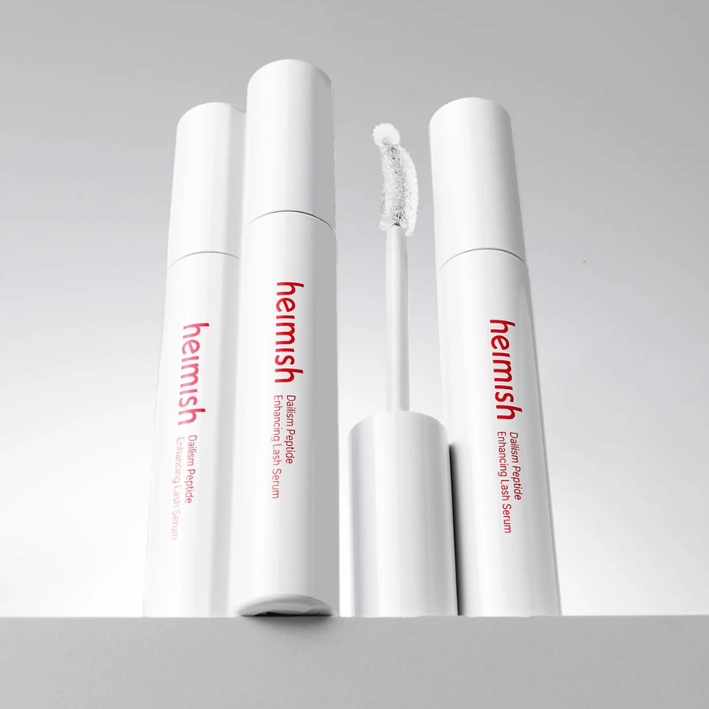 Meerdere verpakkingen van Heimish Dailism Peptide Enhancing Lash Serum worden samen met de gebogen applicatorborstel getoond. De afbeelding benadrukt het elegante ontwerp van het product en de precisieborstel voor eenvoudige applicatie.