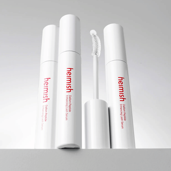 Meerdere verpakkingen van Heimish Dailism Peptide Enhancing Lash Serum worden samen met de gebogen applicatorborstel getoond. De afbeelding benadrukt het elegante ontwerp van het product en de precisieborstel voor eenvoudige applicatie.