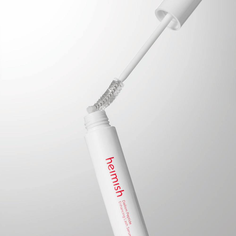 Close-up van de applicator van Heimish Dailism Peptide Enhancing Lash Serum. De borstel wordt uit de witte houder getrokken en toont de transparante serumformule die op de wimpers wordt aangebracht.