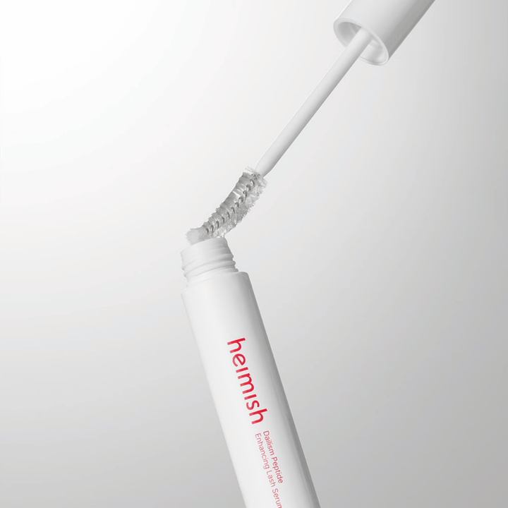 Close-up van de applicator van Heimish Dailism Peptide Enhancing Lash Serum. De borstel wordt uit de witte houder getrokken en toont de transparante serumformule die op de wimpers wordt aangebracht.