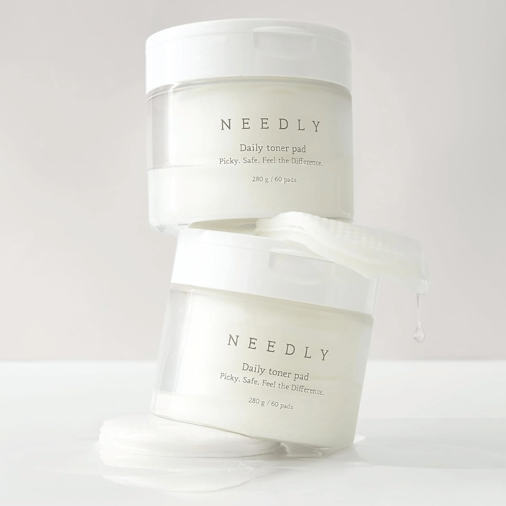 Twee potten met Needly Daily Toner Pad gestapeld op elkaar. De potten zijn wit met minimalistische tekst en bevatten elk 60 toner pads. Een toner pad ligt gedeeltelijk buiten de bovenste pot met vloeistof die eraf druppelt.