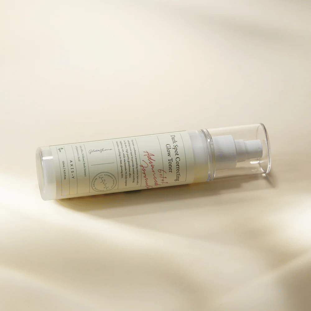 Een liggende fles van "AXIS-Y Dark Spot Correcting Glow Toner" geplaatst op een lichte ondergrond. De fles is doorzichtig met een wit etiket en heeft een witte spraypomp. Het etiket toont informatie over de verhelderende en hydraterende eigenschappen van het product en bevat sleutelingrediënten zoals Glutathion en Niacinamide.