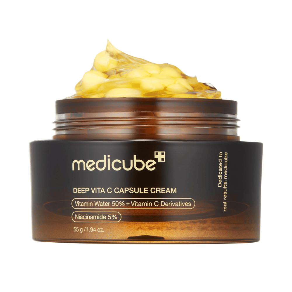 Een close-up van "Medicube Deep Vita C Capsule Cream", die de consistentie van de crème en de capsules van vitamine C laat zien. De verpakking is donkerbruin met gouden tekst en beschrijft de inhoud van vitaminewater (50%) en niacinamide (5%), wat een visuele presentatie geeft van de vochtinbrengende en huidverbeterende eigenschappen van het product.