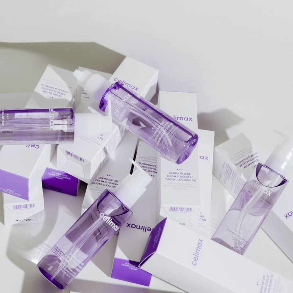 Celimax Derma Nature Fresh Blackhead Jojoba Cleansing Oil in paarse flessen geplaatst tussen witte productverpakkingen met paarse details.