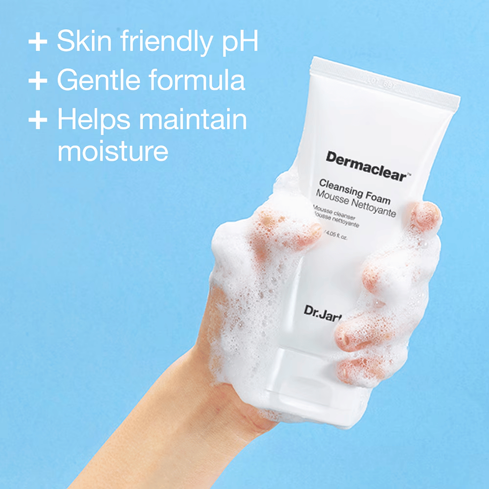 Hand bedekt met schuim houdt Dr.Jart+ Dermaclear Cleansing Foam tegen een blauwe achtergrond, met tekst die huidvriendelijke pH, milde formule en vochtinbrengende eigenschappen benadrukt.