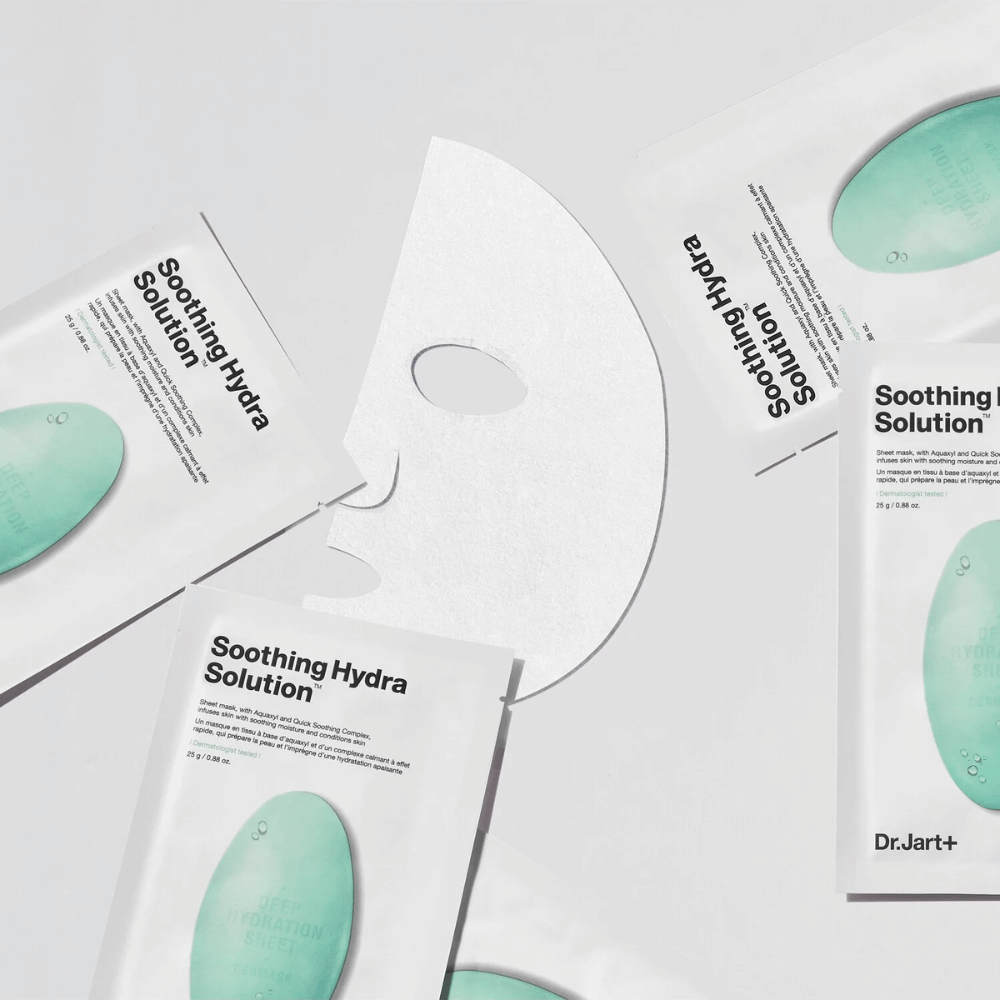 Meerdere verpakkingen van "Soothing Hydra Solution" gezichtsmaskers van Dr.Jart+ liggen verspreid op een lichte ondergrond. Een uitgeklapt masker is in het midden geplaatst, waardoor de witte en geperforeerde structuur zichtbaar is.