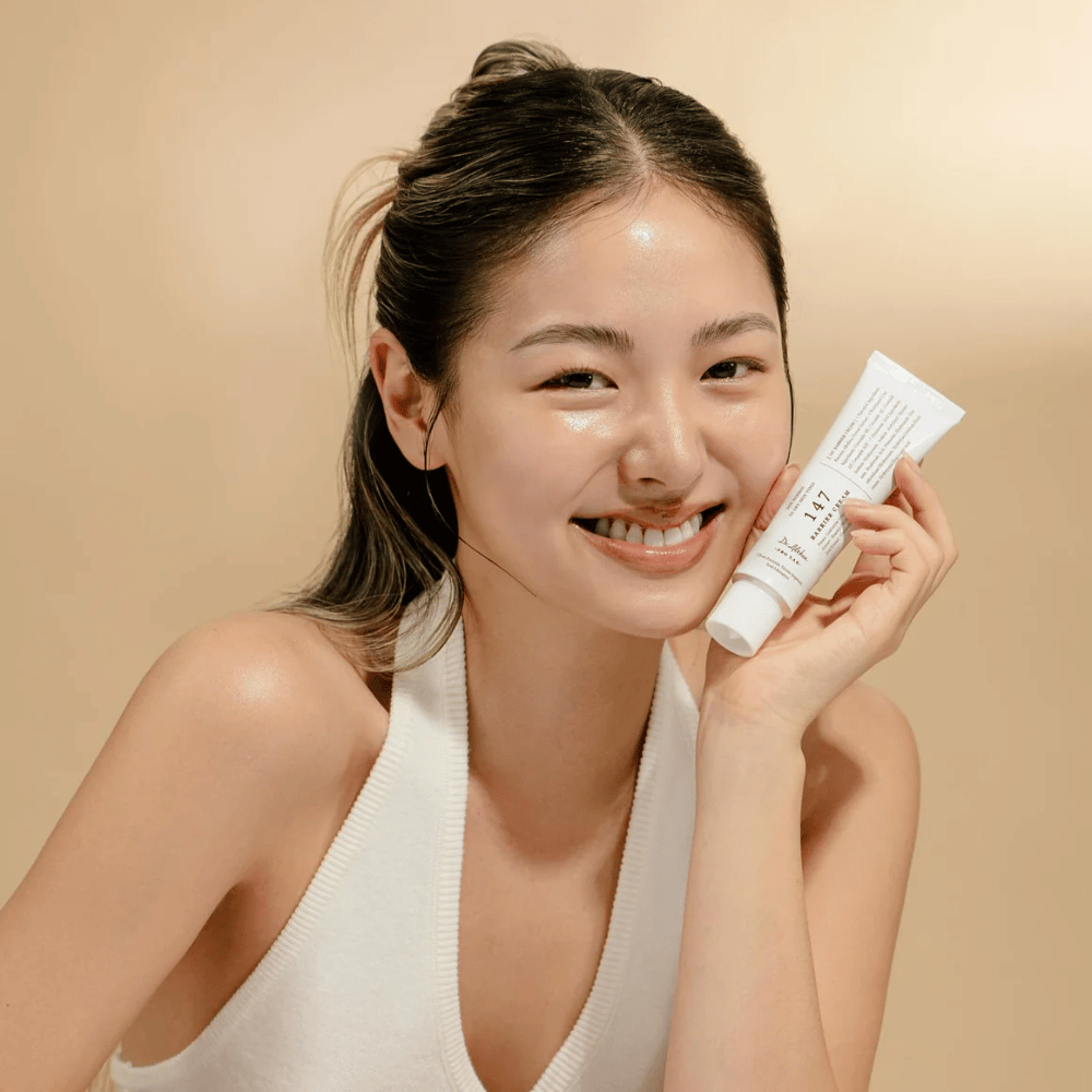 Model met stralende huid die glimlacht en Dr. Althea 147 Barrier Cream vasthoudt – een Koreaanse gezichtscrème voor huidbarrière en hydratatie.