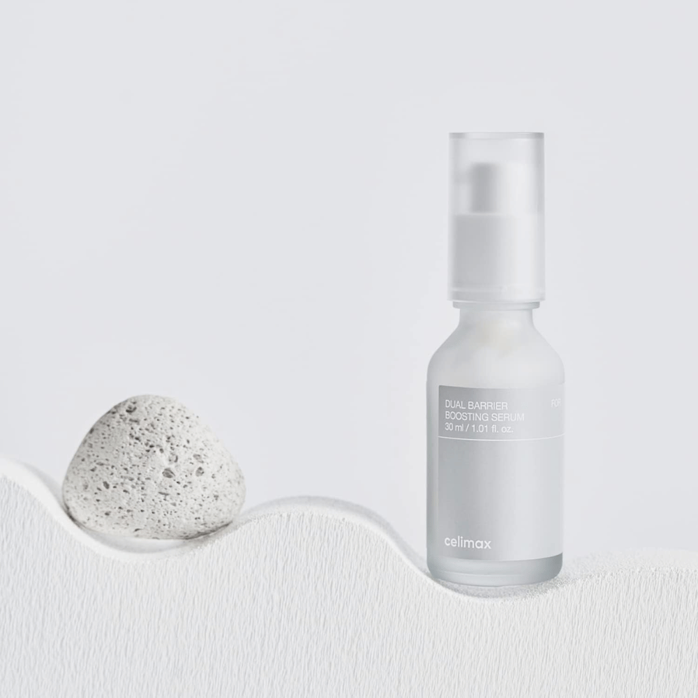 Celimax Dual Barrier Boosting Serum geplaatst op textiel met steen, kalme productafbeelding die hydratatie en versterkte huidbarrière signaleert.