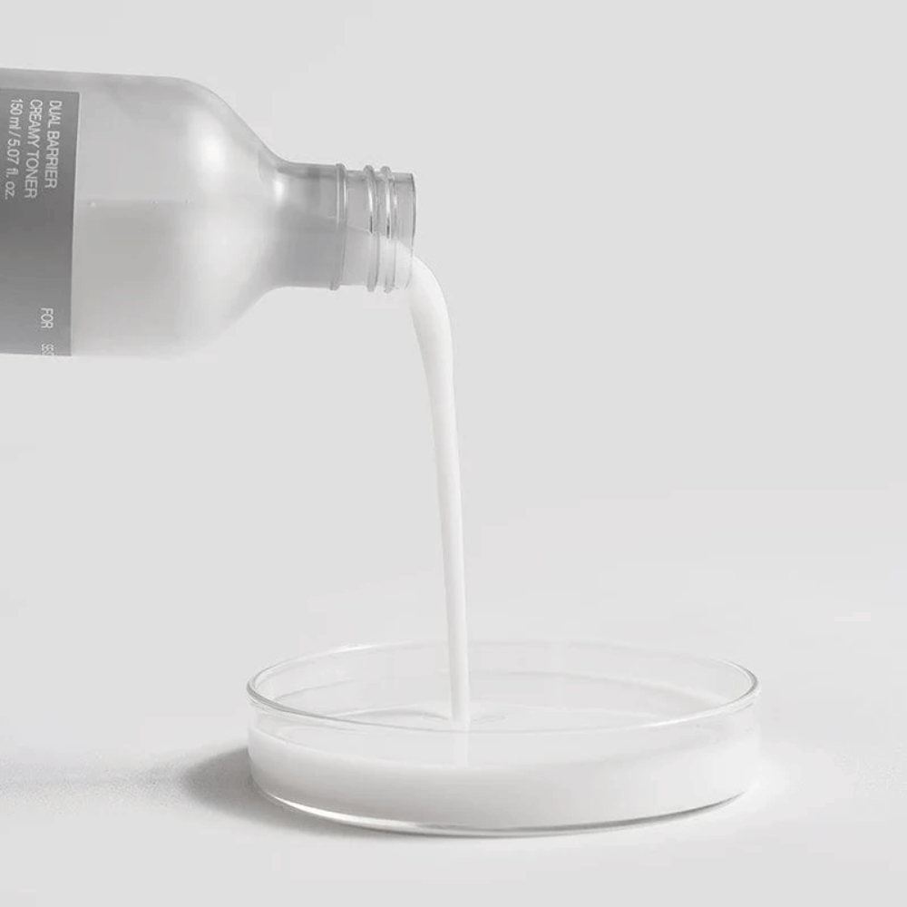 Celimax Dual Barrier Creamy Toner wordt voorzichtig uit de fles in een ronde laboratoriumput gegoten, met de melkachtige en voedzame formule duidelijk in de schijnwerpers.
