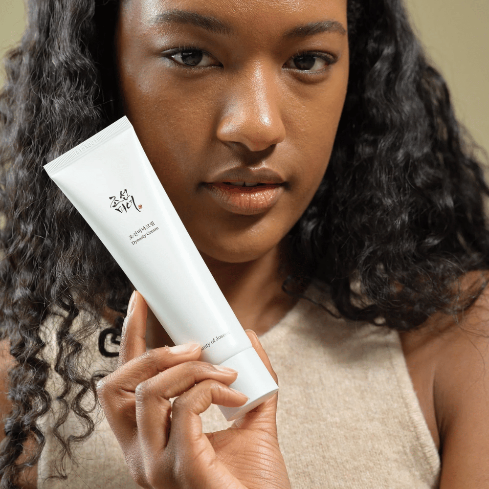 Een vrouw met krullend haar houdt een tube "Beauty of Joseon Dynasty Cream" dicht bij haar gezicht, wat het product benadrukt. De crème, gepresenteerd in een stijlvolle witte tube, is ontworpen om de huid te voeden en te verjongen, wat de aantrekkelijkheid voor verschillende huidtypen benadrukt.