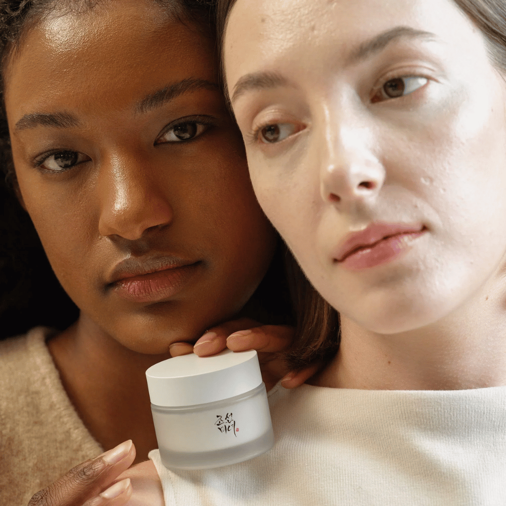 Twee vrouwen, een met een donkerdere en een met een lichtere huidtint, staan dicht bij elkaar met de focus op hun gezichten en een huidcrème van "Beauty of Joseon". De crème is voor hen geplaatst, wat de nadruk legt op het gebruik voor verschillende huidtypen en het vermogen om glans en voeding te geven.