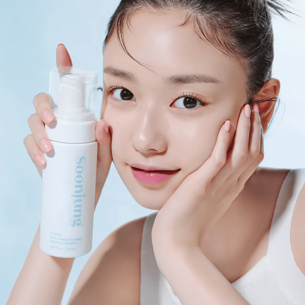 Een jonge vrouw met een heldere huid houdt een witte pompflacon met de tekst "Soon Jung pH 6.5 Whip Cleanser" van ETUDE. Ze lacht naar de camera met een lichte make-up en een frisse uitstraling tegen een lichtblauwe achtergrond.