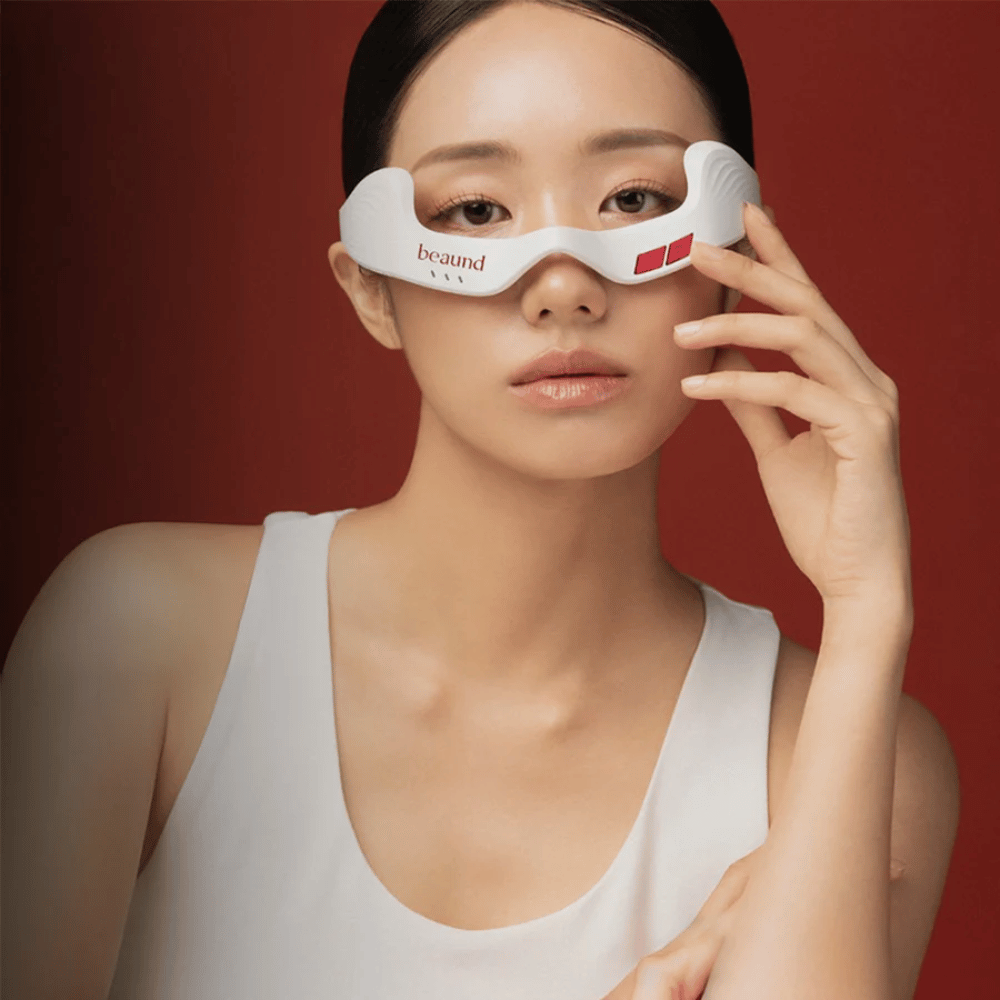 Modelafbeelding waarop een persoon Beaund Eye Rejuv gebruikt, een futuristisch LED-apparaat voor het gebied rond de ogen, tegen een elegante rode achtergrond.
