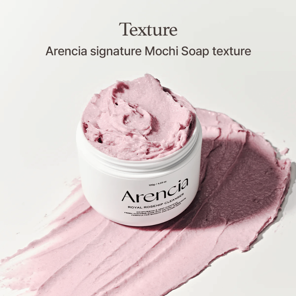 Pot met Arencia Royal Rosehip Cleanser zichtbaar bovenop een uitgesmeerde roze reiniging met mochi-achtige textuur. De tekst erboven beschrijft het product als Arencia's kenmerkende textuur.
