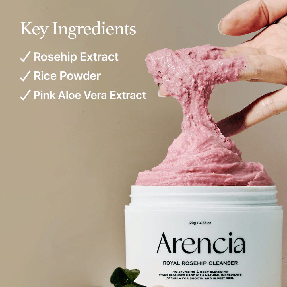 Een hand tilt de roze, luchtige reinigingstextuur uit de pot Arencia Royal Rosehip Cleanser, met een ingrediëntenlijst aan de linkerkant: rozenbottel extract, rijstpoeder en roze aloë vera-extract.