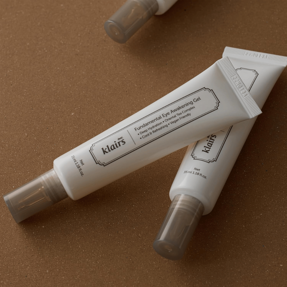 Lifestylebeeld met twee tubes van Klairs Fundamental Eye Awakening Gel geplaatst tegen een warme bruine achtergrond. De compositie benadrukt het elegante ontwerp en de premium uitstraling van het product, terwijl de focus ligt op oogverzorging en hydratatie.