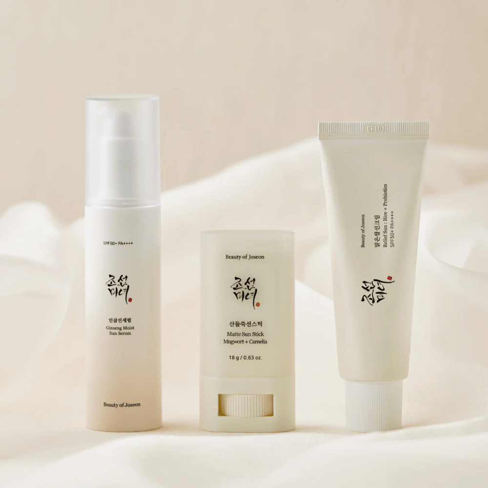 Drie zonnebrandproducten van Beauty of Joseon – Ginseng Moist Sun Serum, Matte Sun Stick en Relief Sun Rice + Probiotics – staan opgesteld op een crèmekleurige stof met zacht licht.