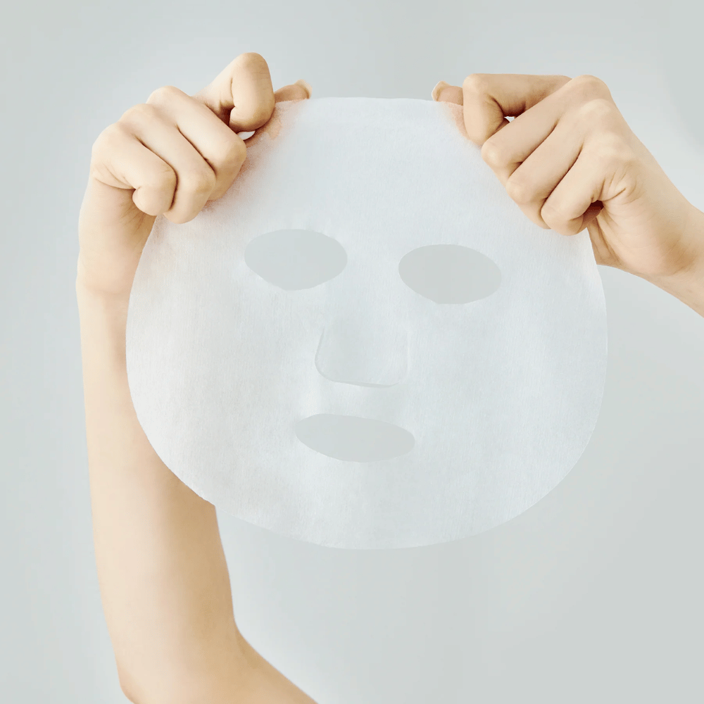 Twee handen houden een wit velmasker omhoog tegen een lichtgrijze achtergrond, met zichtbare uitsparingen voor ogen, neus en mond.
