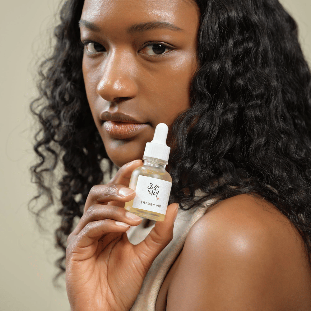 Vrouw met krullend haar houdt een fles Beauty of Joseon Glow Serum met pipet omhoog, een hydraterend en glansgevend huidverzorgingsproduct.