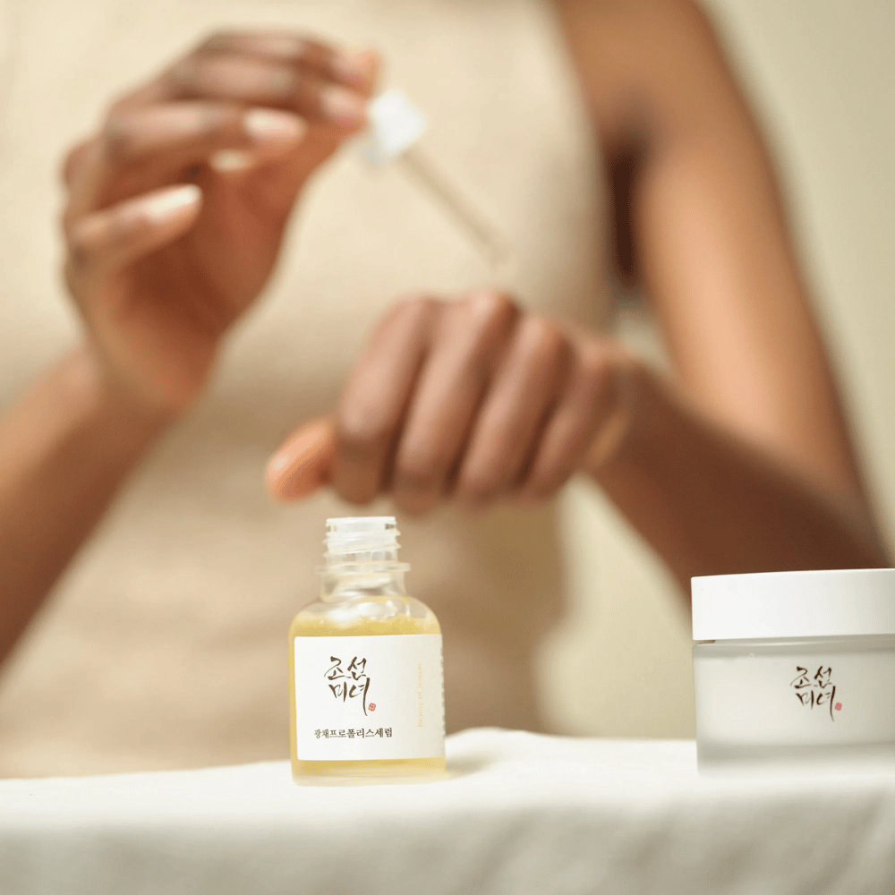 Persoon die Beauty of Joseon Glow Serum met pipet uit glazen fles aanbrengt, met een pot uit dezelfde serie ernaast op tafel.