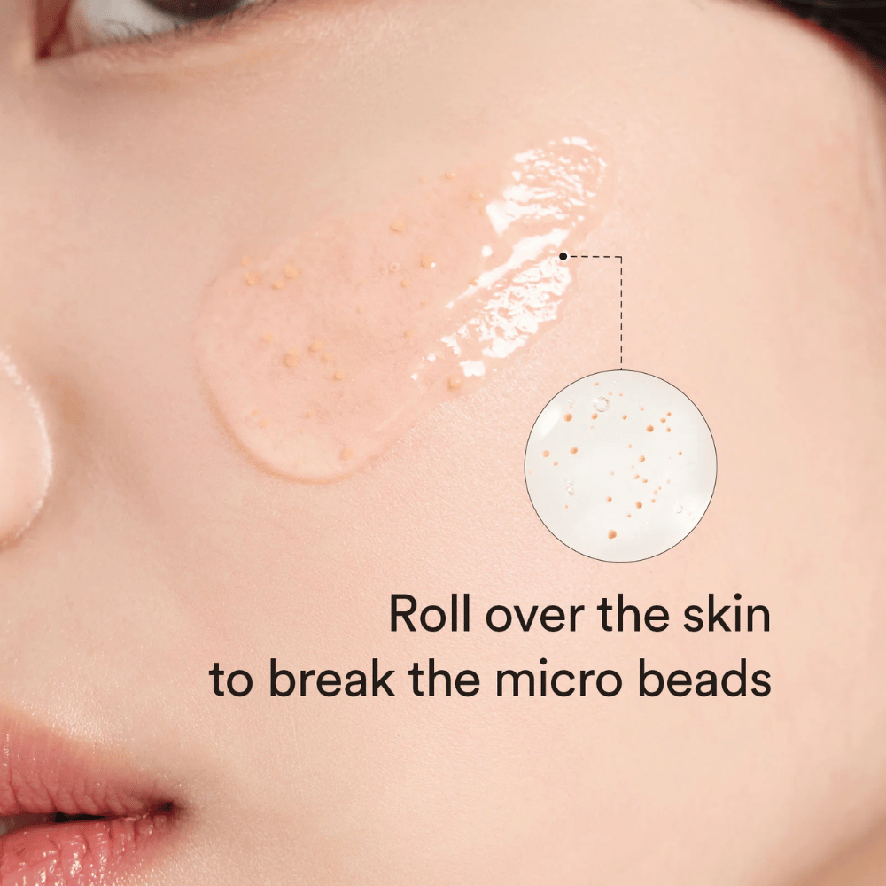 Close-up van een wang met een serum dat kleine microparels bevat, aangebracht op de huid. Een cirkelzoom toont de parels duidelijker. De tekst zegt "Rol over de huid om de microbolletjes te breken".