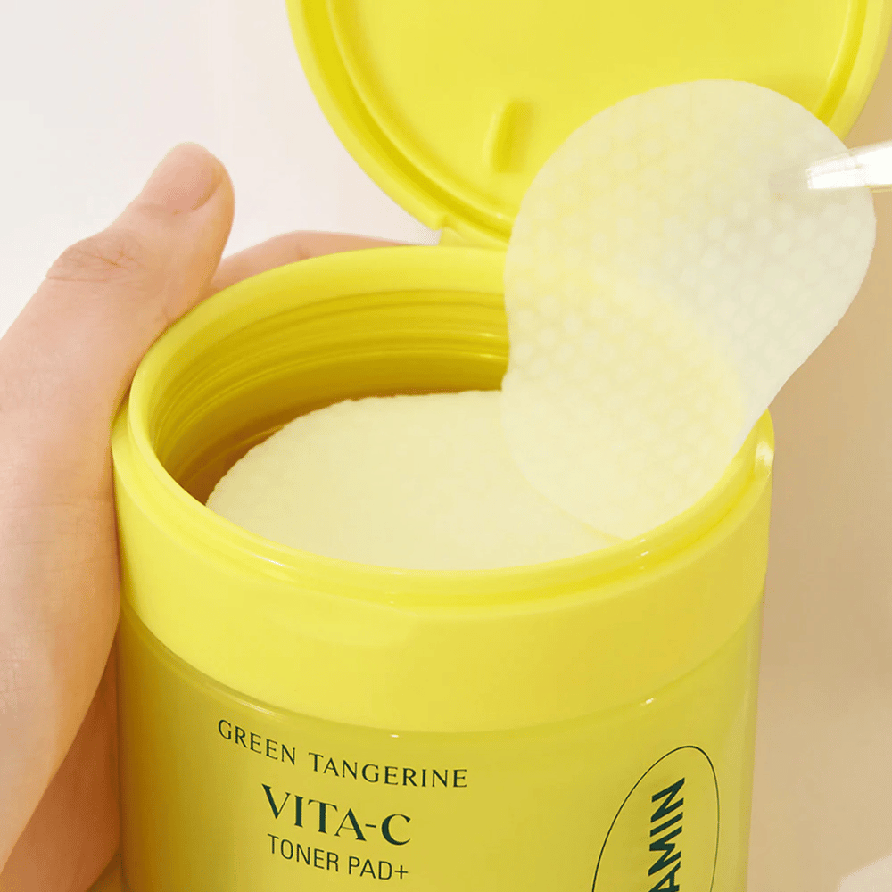 Een hand gebruikt een plastic pincet om een gestructureerd tonerpad op te pakken uit een lichtgele container gemarkeerd "GREEN TANGERINE VITA-C TONER PAD+" van het merk "gooda