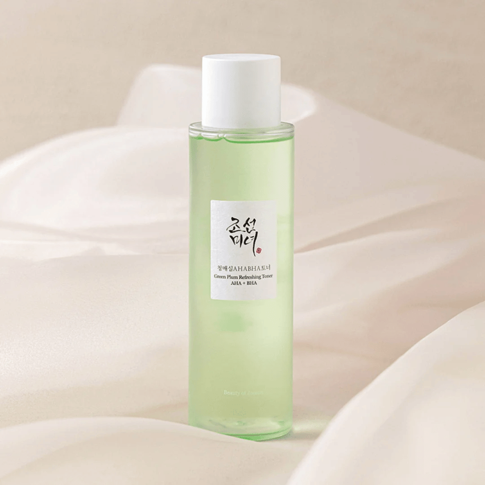 Een fles met "Green Plum Refreshing Toner" AHA + BHA staat op een zachte, lichte achtergrond. Het product heeft een groene vloeistof die frisheid en natuurlijkheid weerspiegelt. Het etiket is wit met zwarte en rode accenten, wat een elegante en strakke look geeft.