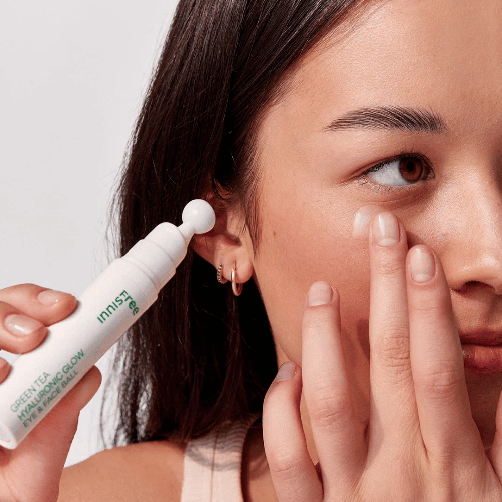 Een persoon brengt Innisfree Green Tea Hyaluronic Glow Eye & Face Ball aan op de huid onder de ogen. Het product heeft een witte, ronde applicator en zit in een witte fles met groene tekst. Het gezicht van de gebruiker is gedeeltelijk zichtbaar en richt zich op het aanbrengen van het product.
