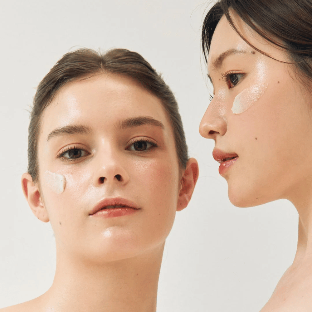 Twee vrouwen met gezichtsmaskers aangebracht op delen van hun gezicht. De ene vrouw heeft een masker op de wang en neus, terwijl de andere het onder het oog en op de wang heeft. Ze hebben natuurlijke make-up en zien er fris uit, wat de hydraterende en verhelderende werking van de maskers benadrukt.