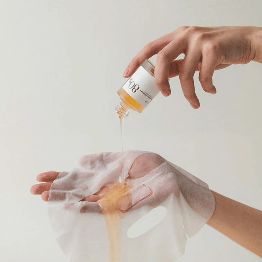 Een hand giet een gouden, dikke vloeistof uit een klein flesje gemarkeerd '108' op een doorzichtig gezichtsmasker dat in een andere hand wordt gehouden. De achtergrond is neutraal en uniform om het product te benadrukken.